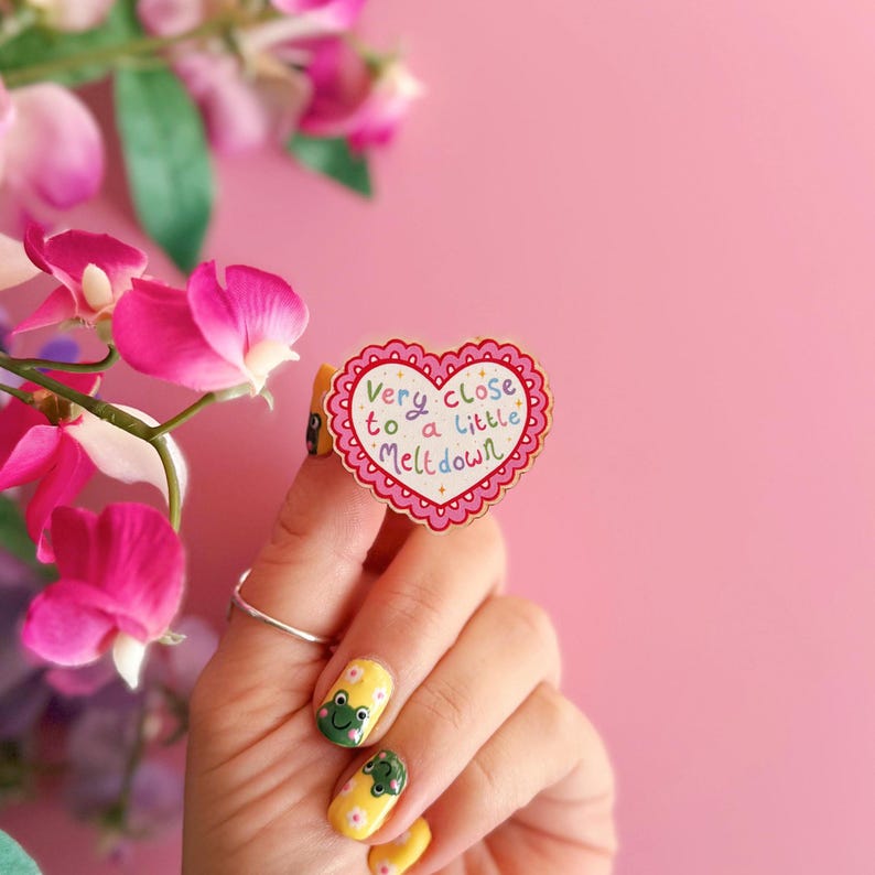Pode incluir: Um alfinete em forma de cora&ccedil;&atilde;o com uma borda ondulada e o texto "Very close to a little meltdown" em letras coloridas. O alfinete &eacute; rosa e branco, e &eacute; segurado por uma m&atilde;o com esmalte de unha amarelo e desenhos de sapos. Flores cor-de-rosa no fundo.