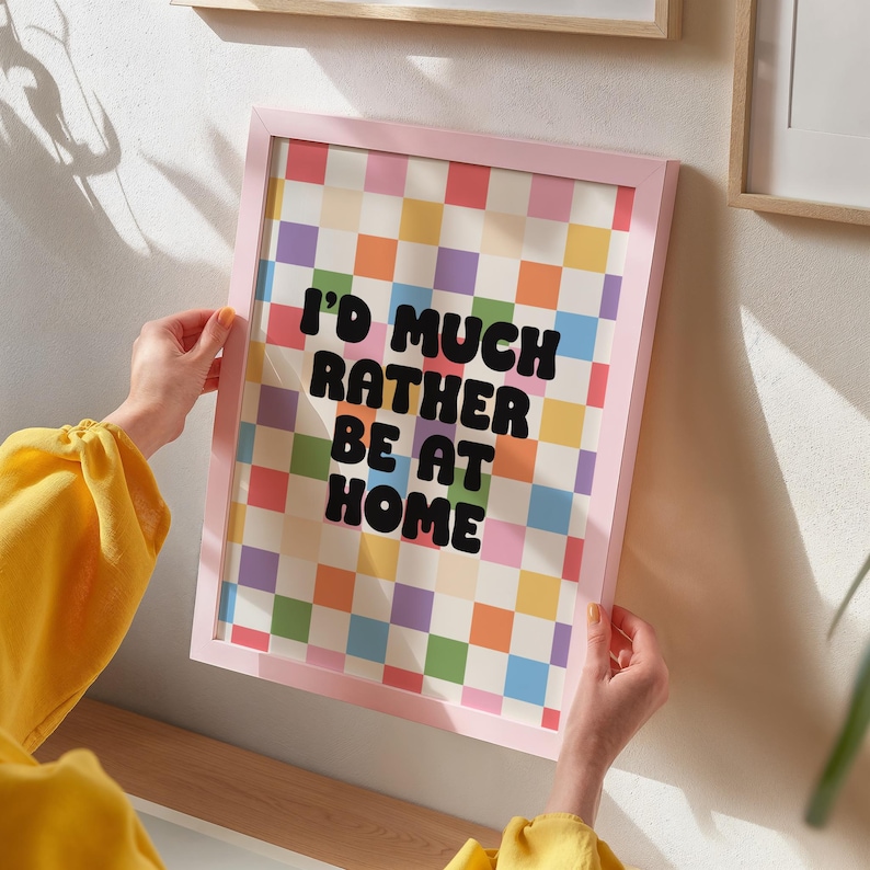 Puede incluir: Una impresi&oacute;n enmarcada en rosa con un fondo a cuadros de colores y el texto "I'd much rather be at home" en negro.