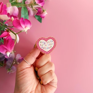 Pode incluir: Um broche em forma de cora&ccedil;&atilde;o com o texto "Very close to a little Meltdown" em letras coloridas. O broche tem uma borda ondulada e uma borda rosa. O fundo &eacute; rosa s&oacute;lido, com flores rosa e roxas no canto superior esquerdo.