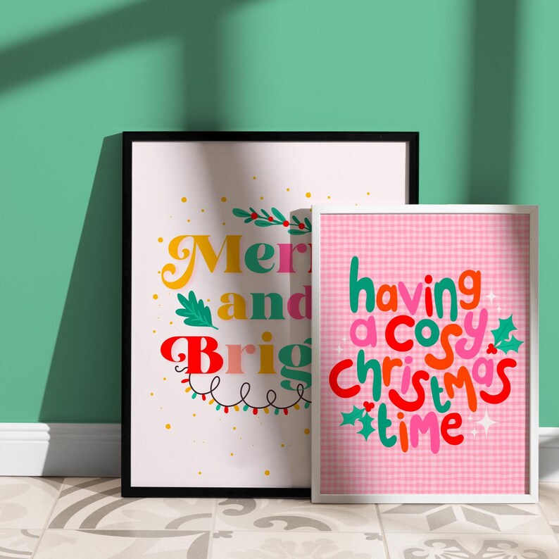 May include: Two framed wall art prints. The larger print features the text Merry and Bright in colorful, stylized lettering with green leaves and string lights. The smaller print has a pink gingham background with the text having a cosy christmas time.