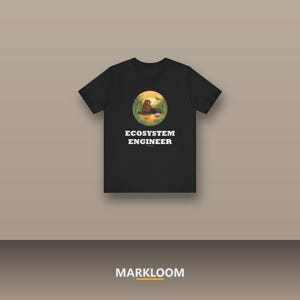 Może przedstawiać: Czarny t-shirt z krótkim rękawem z okrągłą grafiką przedstawiającą bobra w stawie z trzcinami i ważką. Poniżej grafiki znajduje się napis "ECOSYSTEM ENGINEER" białymi wielkimi literami. Koszulka posiada okrągły dekolt.