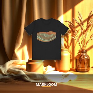 May include: A black short-sleeved t-shirt featuring a central graphic design of horizontal, wavy, layered bands in shades of muted orange, teal, and beige. The shirt is displayed against a warm, sunlit background with fabric and dried plants.