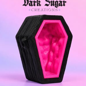 May include: A black coffin-shaped container with a textured exterior and a bright neon pink interior. The text Dark Sugar CREATIONS is printed in black above the object. The container features a small circular opening on the side.