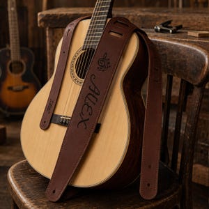 Puede incluir: Una correa de guitarra de cuero marrón oscuro con el nombre Alex y un símbolo de nota musical grabado en la superficie. La correa está colocada sobre una guitarra acústica con cuerpo de madera clara y una roseta de boca circular oscura.