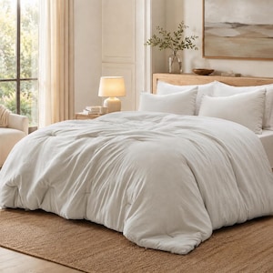 May include: A white tufted comforter set featuring a textured, quilted pattern with subtle indentations across the surface. The bedding includes two matching white pillows placed against a wooden headboard in a bright, neutral-toned bedroom.