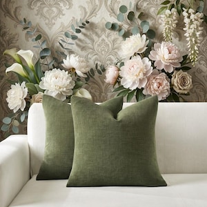 May include: Two square throw pillows in a solid olive green color with a visible woven fabric texture. The pillows are positioned side by side, featuring a simple design with clean edges and a uniform appearance across the front surfaces.