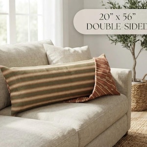 May include: A rectangular lumbar pillow measuring 20 inches by 56 inches, featuring a double-sided design. One side displays horizontal stripes in muted green, brown, and cream, while the other side shows a rust-colored fabric with a subtle pattern.