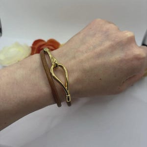 May include: A bracelet featuring a thin brown leather cord wrapped twice around the wrist, secured by a polished gold-tone metal clasp shaped like an elongated teardrop or stylized loop, creating a sleek and simple accessory design.