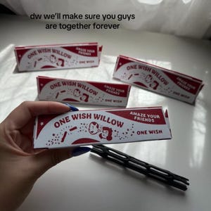 May include: Three rectangular white and red boxes labeled One Wish Willow with the text Amaze Your Friends and One Wish. The boxes feature a graphic of two cartoon faces and stars. A black plastic tool with a grid pattern lies on the surface.