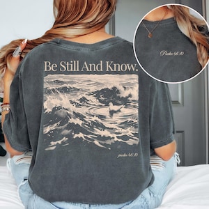 May include: A charcoal grey t-shirt featuring a back graphic of a bird floating on ocean waves with the text Be Still And Know and Psalm 46:10. The front chest area displays the text Psalm 46:10 in a small, light-coloured font.
