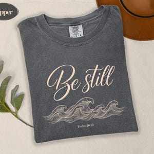 May include: A charcoal grey t-shirt with a crew neckline and a Comfort Colors label. The front features the light pink text 'Be still' in a cursive script above a line drawing of ocean waves and the small text 'Psalm 46:10' at the bottom.