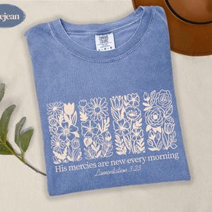 Puede incluir: Una camiseta de color azul jean con un gráfico de líneas blancas de cuatro paneles florales verticales. Debajo de las flores, el texto dice: His mercies are new every morning, Lamentation 3:23. La camiseta tiene cuello redondo y etiqueta Comfort Colors.