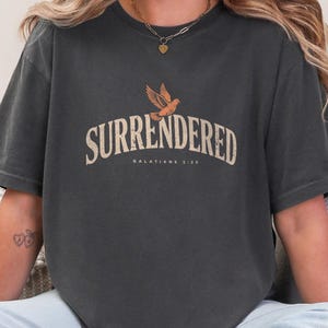 Puede incluir: Una camiseta de manga corta gris carbón que presenta la palabra SURRENDERED en una fuente beige desgastada, situada bajo un pequeño gráfico de un pájaro volando. Debajo del texto principal, se lee GALATIANS 2:20 en color beige.