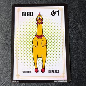 Könnte beinhalten: Eine rechteckige Spielkarte mit einer Illustration eines gelben Gummihuhns mit roter Fliege und roten Füßen. Die Karte zeigt den Text BIRD, die Zahl 1 mit einem Schildsymbol, TOKEN UNIT, DEFLECT, MEM T03 und RUNETERRABOUND auf weißem Grund.