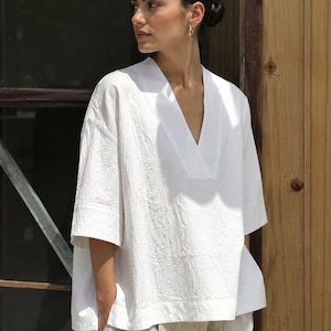 May include: A white short-sleeved top with a deep V-neckline and a textured fabric finish. The garment features a loose, boxy silhouette with wide sleeves and a straight hemline, paired with light beige trousers that have a visible pleated detail.