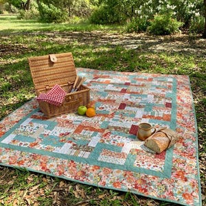 May include: A rectangular patchwork quilt featuring a floral and geometric pattern in shades of teal, orange, red, and white. The quilt is laid out with a woven wicker picnic basket, a red and white checkered napkin, two pieces of fruit, a mug, and bread.