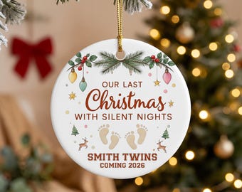 May include: A round white ceramic ornament hanging by a gold string. It features green pine branches, red berries, and colorful ornaments. Text reads: Our Last Christmas With Silent Nights, Smith Twins Coming 2026, with baby footprints.