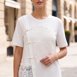 May include: A white short-sleeved top featuring a round neckline and an asymmetrical front panel. The garment is adorned with a large, circular white lace fan appliqué and three decorative white toggle closures. A wide lace trim borders the lower hem.