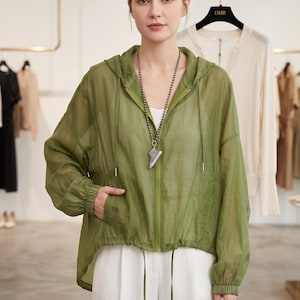 May include: A sheer olive green hooded jacket with a front zipper closure, long sleeves with elastic cuffs, and a drawstring hem. The jacket is worn over a white top and white trousers, paired with a long beaded necklace featuring a sneaker pendant.