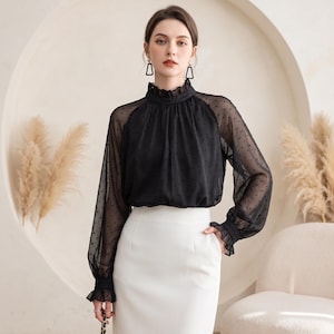 Op de afbeelding: Een zwarte blouse met lange mouwen van transparante stof met een subtiel stippenpatroon, een hoge kraag met ruches en geplooide manchetten met ruches, gecombineerd met een effen witte rok met hoge taille.