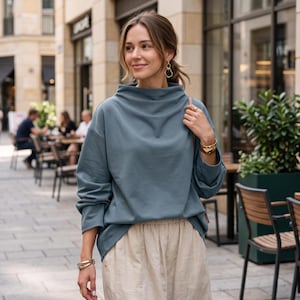 May include: A slate blue long-sleeve sweatshirt with a high, draped cowl neckline and dropped shoulders, paired with loose-fitting beige trousers featuring an elasticized waistband. The outfit is accessorized with gold hoop earrings and bracelets.