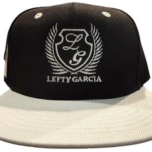 Pode incluir: Um boné de beisebol preto com aba reta cinza-clara. A parte frontal apresenta um bordado branco de um escudo contendo as letras LG, ladeado por duas asas, com o texto LEFTY GARCIA bordado em letras maiúsculas brancas abaixo do escudo.