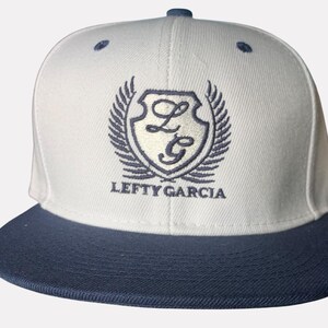 Pode incluir: Um boné de beisebol branco com aba reta azul-escura e ilhós azul-escuro. A frente apresenta um logótipo de escudo azul-escuro bordado com as letras LG, ladeado por duas asas, com o texto LEFTY GARCIA bordado por baixo do escudo.