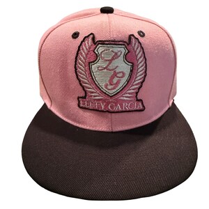 Pode incluir: Um boné de beisebol rosa com aba reta marrom escura e um botão superior preto. A frente apresenta um brasão bordado com asas, as letras LG e o texto LUFFY GARCIA em fio rosa e branco sobre um fundo de tecido rosa claro.