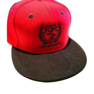 Pode incluir: Um boné de beisebol vermelho brilhante com aba preta reta e um botão preto no topo. A frente apresenta um logótipo de escudo preto bordado com asas e o texto LEFTY GARCIA. O boné tem ilhós pretos para ventilação na coroa.