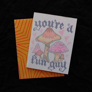 Könnte beinhalten: Eine weiße Grußkarte mit einer farbenfrohen Illustration von vier Pilzen in Rosa- und Orangetönen, umgeben von kleinen rosa Herzen. Der Text "you're a fun guy" ist in einer dunklen gotischen Schrift gedruckt. Ein orangefarbener Umschlag mit Linien liegt dahinter.