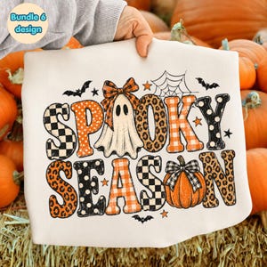 May include: A white t-shirt featuring the text SPOOKY SEASON in large, patterned letters. The design includes a ghost with an orange plaid bow, a pumpkin with a black and white plaid bow, spiderwebs, bats, and stars in orange and black.