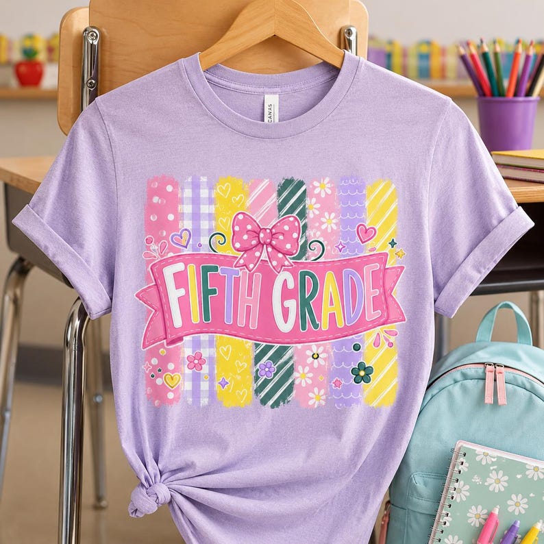 May include: A light purple t-shirt featuring a graphic print with vertical stripes in various colors and patterns, a pink banner with the text FIFTH GRADE, and a pink polka-dot bow. The design includes small hearts, flowers, and stars.