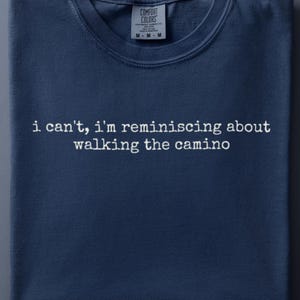 May include: A navy blue crew neck t-shirt featuring the white text "i can't, i'm reminiscing about walking the camino" printed in a typewriter-style font across the chest. The shirt includes a Comfort Colors brand label at the inner neckline.