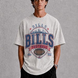 Op de afbeelding: Een lichtgrijs t-shirt met korte mouwen en een grafische print met de tekst BILLS, FOOTBALL, EST. 1960 en BUFFALO, USA. Het ontwerp bevat een stadssilhouet, een illustratie van een football en rode en blauwe accenten op de voorzijde.