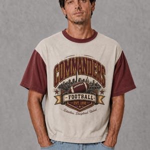 May include: A short-sleeve t-shirt with a cream-coloured body and maroon sleeves and collar. The graphic features the text COMMANDERS, a football, a city skyline, and the text FOOTBALL EST. 1932, Relentless. Disciplined. United. in maroon and gold.