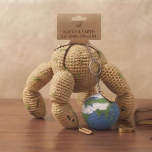 May include: A crochet bundle featuring a tan, multi-legged creature and a small globe keychain. The packaging header reads ROCKY & EARTH 2-IN-1 BUNDLE KEYCHAIN SET. The tan creature has green accents, and the globe features blue, green, and white yarn.