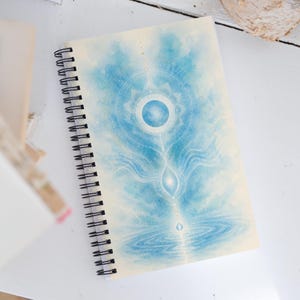 Può includere: Un taccuino a spirale con un'illustrazione ad acquerello nei toni del blu e del bianco. L'opera raffigura un globo centrale luminoso, motivi circolari geometrici e linee fluide che scendono verso una superficie d'acqua increspata.