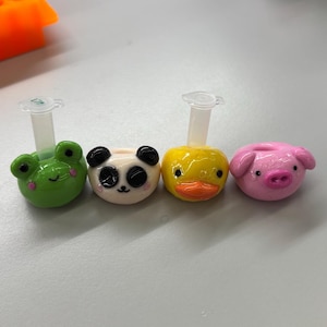 May include: Four small, decorative animal-shaped toppers for laboratory tubes. The set includes a green frog, a black and white panda, a yellow duck, and a pink pig. Each figure features a small, clear plastic tube attachment on the top.