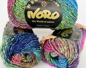 May include: Three skeins of Noro yarn featuring a variegated blend of blue, green, pink, and yellow fibers. Each skein has a black paper label with the text Noro, the World of nature, Made in Japan, and Japanese characters printed in gold.
