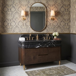Front view of a luxury master bathroom centered around a handcrafted Nero Marquina double marble vanity with dual integrated sinks, elegant molded stone countertop, brushed brass faucets, rich dark walnut cabinet, oval mirror, decorative wallpaper, and designer lighting for a refined contemporary classic interior.