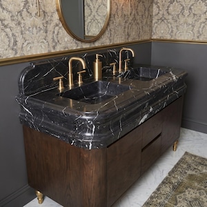 Angled view of a handcrafted Nero Marquina marble double vanity showcasing dual integrated stone basins, decorative molded marble edge profile, matching backsplash, brushed brass fixtures, and a dark walnut cabinet. The dramatic black marble with bold white veining creates a timeless luxury bathroom centerpiece.