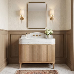 Front view of a luxury bathroom featuring a custom Carrara marble vanity with an integrated sink, fluted natural oak cabinet, brushed brass wall-mounted faucet, designer mirror, elegant sconces, decorative hydrangeas, and genuine white marble with graceful gray veining. A handcrafted statement vanity designed for premium residential and boutique hospitality interiors.