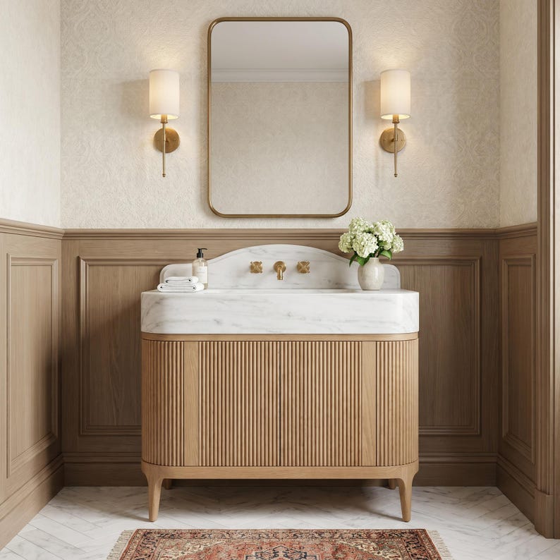 Front view of a luxury bathroom featuring a custom Carrara marble vanity with an integrated sink, fluted natural oak cabinet, brushed brass wall-mounted faucet, designer mirror, elegant sconces, decorative hydrangeas, and genuine white marble with graceful gray veining. A handcrafted statement vanity designed for premium residential and boutique hospitality interiors.