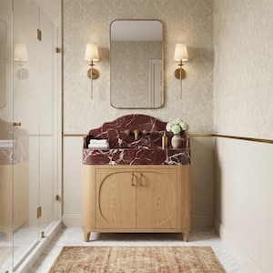 May include: A bathroom vanity featuring a deep red marble countertop with white veining, a brass faucet, and a light wood cabinet with two doors. Above the vanity is a rectangular mirror flanked by two brass wall sconces with white shades.
