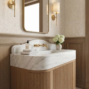 Side perspective of a handcrafted Carrara marble bathroom vanity with an integrated marble sink, rounded waterfall edges, sculpted backsplash, brushed brass fixtures, and fluted natural oak cabinetry. Genuine white marble with soft gray veining creates a refined centerpiece suited for modern, classic, and transitional bathroom designs.