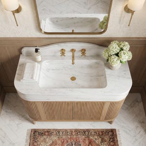 Top view of a custom Carrara marble bathroom vanity showcasing an integrated marble basin, rounded countertop edges, sculpted backsplash, brushed brass faucet and drain, fluted natural oak cabinetry, folded towels, decorative flowers, and authentic white marble with delicate gray veining. Designed for elegant transitional and contemporary luxury bathrooms.