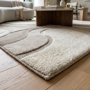 Low angle view of a cream and beige abstract wave area rug highlighting the sculpted high-low surface, raised organic curves, soft neutral colors and clean finished edge on a natural wood floor.
