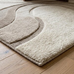 Corner view of a rectangular cream and beige abstract wave area rug showing the clean finished edge, layered organic curves, dimensional surface and sophisticated neutral color palette.