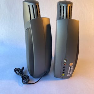 May include: Two dark gray vertical computer speakers with a curved design and a mesh front panel. The back of one speaker features a series of circular input ports, including red, white, and yellow jacks, along with a black power cable attached.