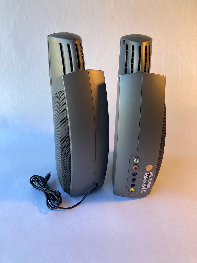 May include: Two dark gray vertical computer speakers with a curved design and a mesh front panel. The back of one speaker features a series of circular input ports, including red, white, and yellow jacks, along with a black power cable attached.